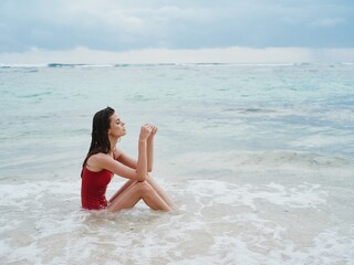 Woman with a beautiful tan tourist in a red swimsuit sitting on the sand on the beach in the ocean in the waves pensive