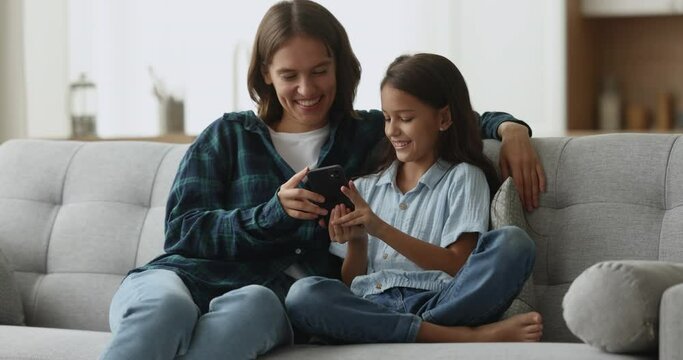 Happy young mom and cheerful daughter kid watching funny online video content on smartphone, sitting on couch at home together, holding mobile phone, looking at screen, laughing