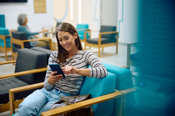 Young happy woman using cell phone in waiting room at doctor's office. - obrazy, fototapety, plakaty