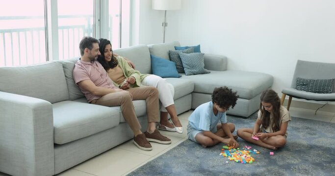 Happy Latin middle aged couple of parents discussing family plans while two kids playing construction game on floor, sitting on comfortable couch in living room, talking, smiling