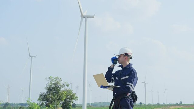 Wide shot technician worker hold laptop and look to the monitor for working also use walkie talkie to contact other in front of windmill or wind turbine with blue skay in area of power plant factory.