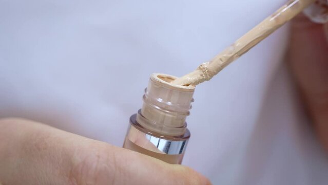 Beige skin concealer pouring from a brush into the bottle in woman hands on light background close up