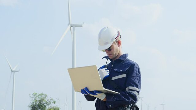 Technician worker hold laptop and look to the monitor for working also use walkie talkie to contact other in front of windmill or wind turbine with blue skay in area of power plant factory.