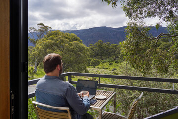 A man working remotely from his balcony looking over bushland and mountains