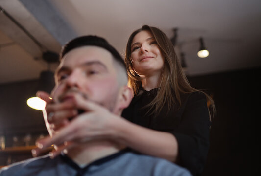 Young long hair female barber gently massaging face skin after bearded man shaving procedure Smiling at mirror. Modern black-style barber shop. Hair and skin care service, local small business concept