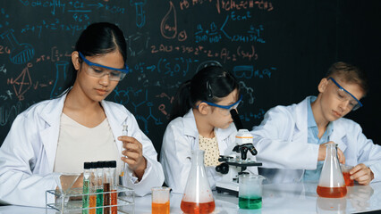 Cute girl looking under microscope while student doing experiment at blackboard with theory...