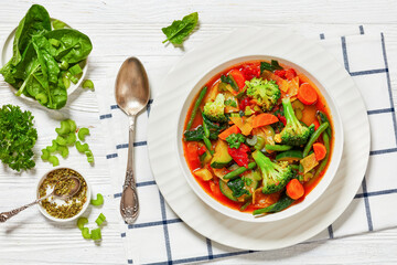 vegetable soup with green beans, broccoli, carrot, celery, spinach, cabbage, zucchini, tomatoes in white bowl on white wooden table with spoon and ingredients, horizontal view, flat lay