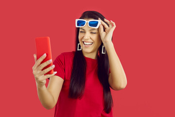 Joyful excited woman uses a mobile phone for online shopping, chatting or browsing social networks. Happy woman raises glasses while looking at modern smartphone while standing on red background