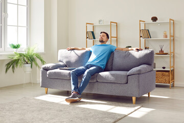 Young relaxed man sitting on comfortable couch in the living room at modern home and having a rest...
