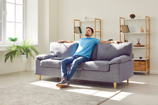 Young relaxed man sitting on comfortable couch in the living room at modern home and having a rest from a work with closed eyes. Calm peaceful guy enjoying quiet time alone in the room and dreaming.