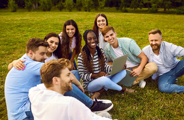 University friends working and studying together using laptop while sitting on grass of campus park. Multiracial men and women in casual clothes sitting on green lawn with laptop chatting and smiling.