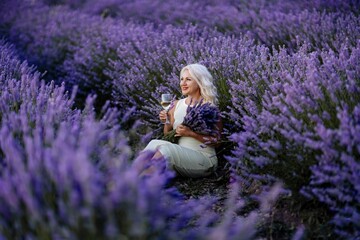 Blonde lavender field holds a glass of white wine in her hands. Happy woman in white dress enjoys...
