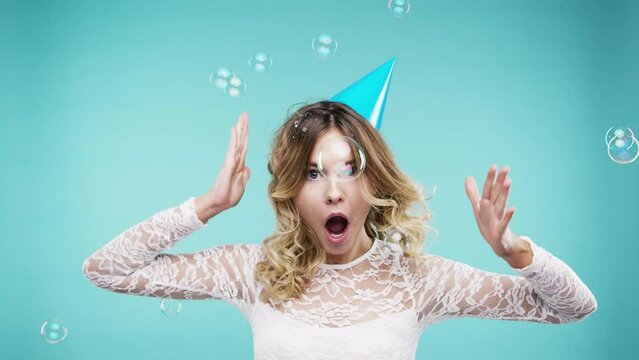 Woman, bubbles and funny face in studio for dance party, celebration and crazy or happy on new year. Silly person, winner or model with energy, playful and funky hat for event on a blue background