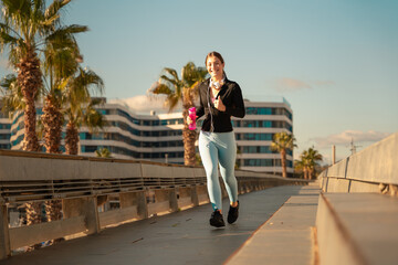 Fototapeta premium A happy fitness woman runs with headphones and water bottle looking at camera. Workout concept and healthy lifestyle.