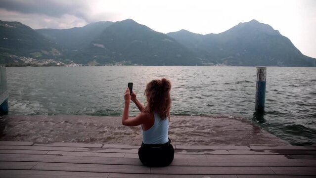 young cute girl with curly hair from behind sitting on a wooden pier on the shores of Lake Iseo on a cloudy and windy summer day. The girl is busy taking photos of her with her phone