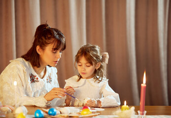 Naklejka premium Cozy moment at home. Beautiful young woman, mother sitting at home with daughter, preparing for Easter, painting eggs. Concept of holiday, Easter, family, motherhood and childhood