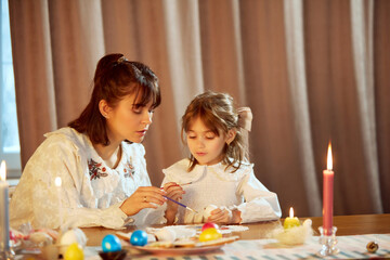 Mother and child painting Easter eggs together at table with lit candle. Concept of holiday, Easter, family, motherhood and childhood. Family bonding activities