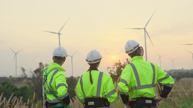 Back of professional technician man and woman workers stand and discuss together about work and look to area of wind turbine or windmill with evening light.