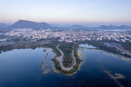 aerial drone shot at dawn dusk with road loop extending into fateh sagar lake with aravalli hills in distance hidden in fog showing cityscape of Udaipur Rajasthan