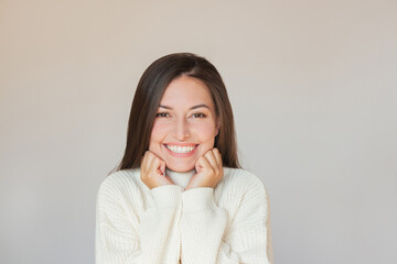 A young pretty Caucasian brunette woman in a beige sweater rests her head on her hands, looks at the camera and smiles beautifully. Portrait of a beautiful woman. Isolated on a beige background