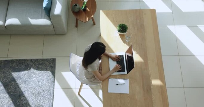 Young Asian woman working on business project at home, typing on laptop, sitting at workplace table with computer and notebook, using Internet technology for communication. Top view