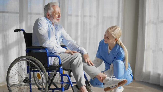 Nurse talking senior man in wheelchair. Female social welfare physical therapist looks after senior male, listening, providing medical care at home. Living with legs paralysis, special needs people.