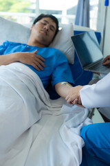 Young Asian male patient lying in bed while checking his pulse. Consult and explain with the nurse who takes notes and assists in the hospital in the recovery room in the hospital department.