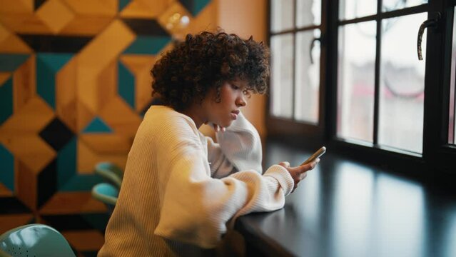 Young woman with phone chatting with friends. African American girl hold in hand use mobile phone in cafe. Teenager spend free spare time in living room with phone. Communication on social networks.