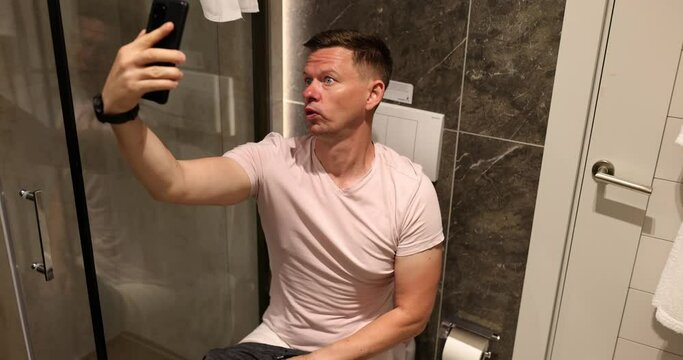 Man grimaces jokes and takes selfie toilet. Fool joking in toilet with smartphone