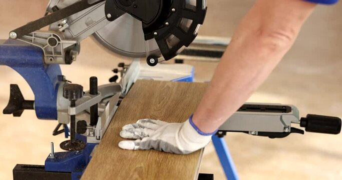Man in protective gloves works with miter saw, cutting wooden board of laminate. Handyman using material handling equipment for home renovation