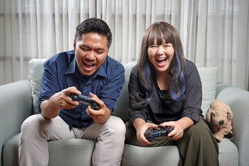 Happy young couple sitting on the sofa in the living room playing video games together having fun. Man and woman playing games on console at home, recreation concept