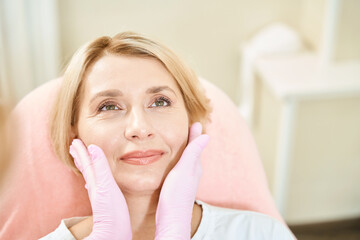 Cropped female cosmetologist hands examining and touching face of smiling woman