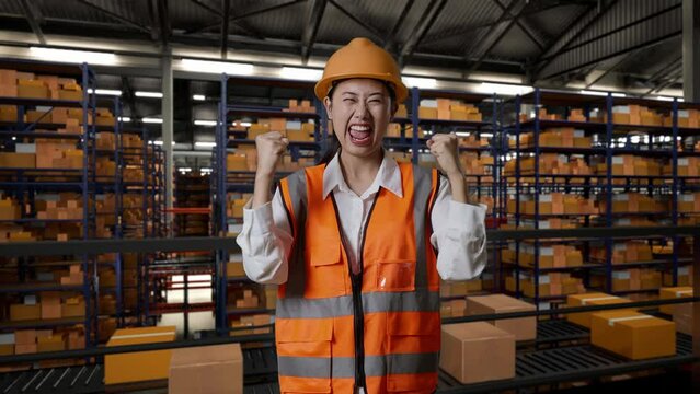 Asian Female Engineer With Safety Helmet Standing And Screaming Goal Celebrating Working In The Warehouse With Shelves Full Of Delivery Goods
