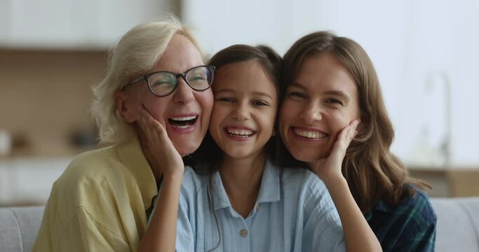 Happy little kid girl, cheerful young mom and elderly grandma sitting close with face touches, looking at camera, smiling, laughing, closing eyes, enjoying closeness, generation connection
