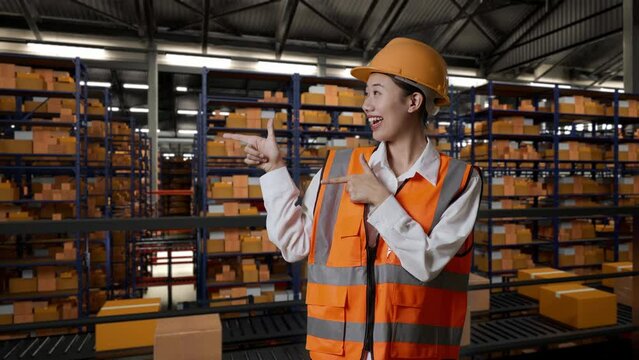 Asian Female Engineer With Safety Helmet Standing In The Warehouse With Shelves Full Of Delivery Goods. Smiling And Pointing To Side Recommends About Something In The Storage
