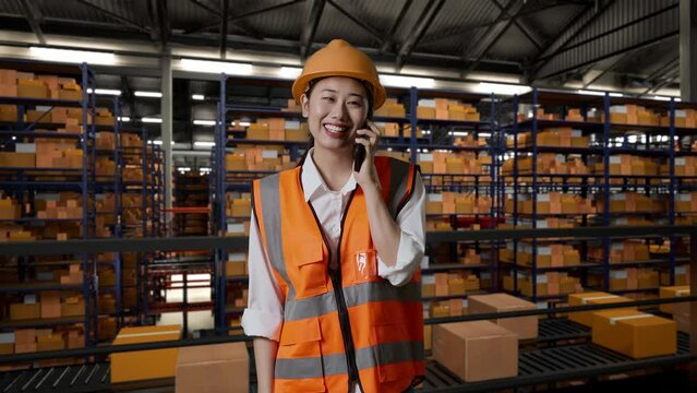 Asian Female Engineer With Safety Helmet Talking On Smartphone While Standing In The Warehouse With Shelves Full Of Delivery Goods
