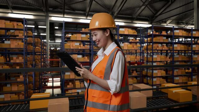 Side View Of Asian Female Engineer With Safety Helmet Working On A Tablet While Standing In The Warehouse With Shelves Full Of Delivery Goods
