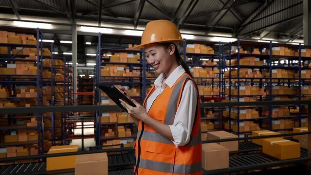 Side View Of Asian Female Engineer With Safety Helmet Standing In The Warehouse With Shelves Full Of Delivery Goods. Taking Note On The Tablet In The Storage
