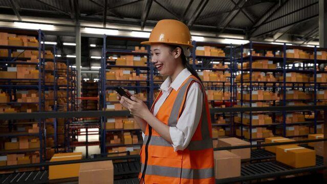 Side View Of Asian Female Engineer With Safety Helmet Using Smartphone While Standing In The Warehouse With Shelves Full Of Delivery Goods
