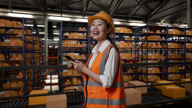 Side View Of Asian Female Engineer With Safety Helmet Using Smartphone And Looking Around While Standing In The Warehouse With Shelves Full Of Delivery Goods
