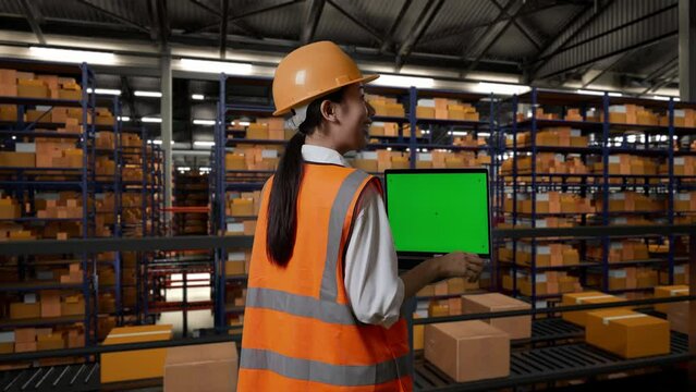 Back View Of Asian Female Engineer With Safety Helmet Standing In The Warehouse With Shelves Full Of Delivery Goods. Looking At Green Screen Laptop And Looking Around The Storage
