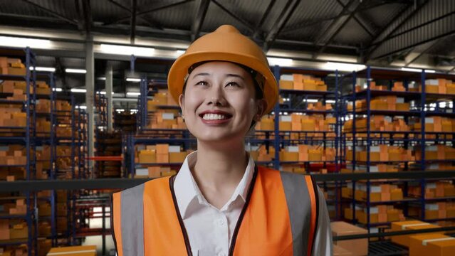 Close Up Of Asian Female Engineer With Safety Helmet Standing In The Warehouse With Shelves Full Of Delivery Goods. Looking Around, Checking The Stock On Racks
