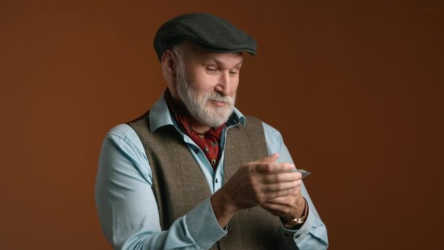 A man of seasoned years expresses curiosity as he counts a handful of bills, attired in a classic flat cap and tweed vest, against a warm brown background. High quality 4k footage