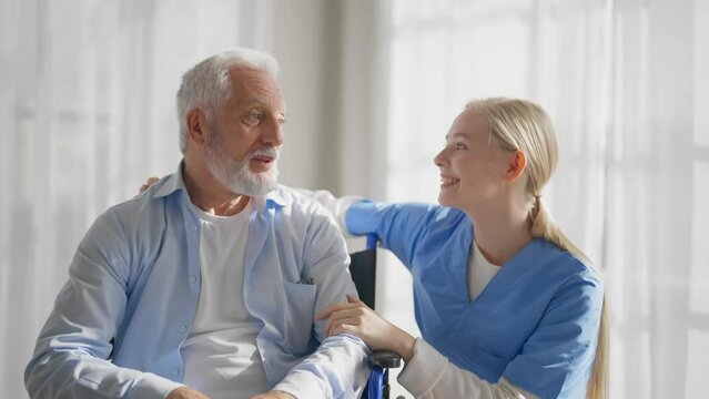 Nurse talking senior man in wheelchair. Woman social welfare physical therapist looks after senior male, listening, providing medical care at clinic. Living with legs paralysis, special needs people.