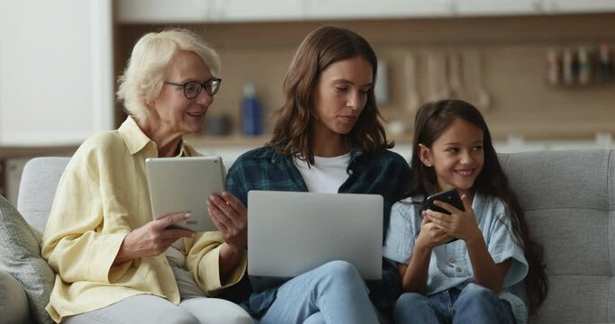 Positive child, mother and grandmother discussing online chat, safe Internet communication for kids, holding computers and smartphone, resting on home sofa together, talking