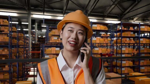 Close Up Of Asian Female Engineer With Safety Helmet Standing In The Warehouse With Shelves Full Of Delivery Goods. Talking On Smartphone And Looking Around The Storage
