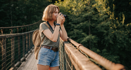 Attractive young female traveler photographing with camera while standing on the scenic bridge
