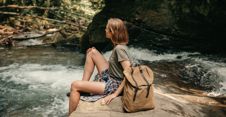 Young female traveler with backpack resting while sitting by the mountain river