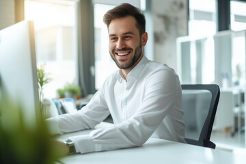 Portrait of a young man with a beard, wearing smart casual attire, displaying a warm, friendly smile as he works at his computer in a modern, naturally lit office space. His appearance is neat and pro - obrazy, fototapety, plakaty