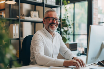 Fototapeta premium Portrait of a cheerful, bearded, and bespectacled older man with white hair, engaging with the camera with a bright smile while working at his computer in a sunny, modern office environment.